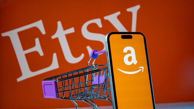 Miniature shopping cart with smartphone showing Amazon app logo on Etsy logo background.