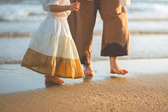 Asian toddler girl and parent walking barefoot on the beach during golden hour sunset. Concept of family travel, childhood memories, and summer vacation. A peaceful moment of a mother and daughter