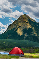 Red camping tent pitched in a green valley beneath a towering mountain peak under a cloudy sky. Ideal for outdoor recreation, travel, and adventure themes. © Christina