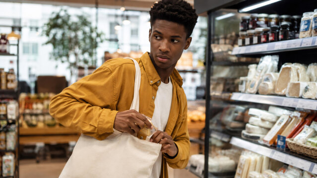 African american man hiding product in bag in grocery store. Shoplifter looking away nervously while stealing food from shelf. Surveillance and retail security monitoring concept.