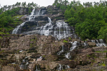 Multi-tiered Norwegian waterfall streaming over dark rock with lush green trees, viewed from below in summer. © Andrei Tomas