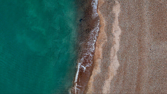 Top-down aerial view of Eastbourne's pebble beach, emerald shoreline and coastal road, revealing striking natural patterns and urban geometry along the East Sussex coast, England.