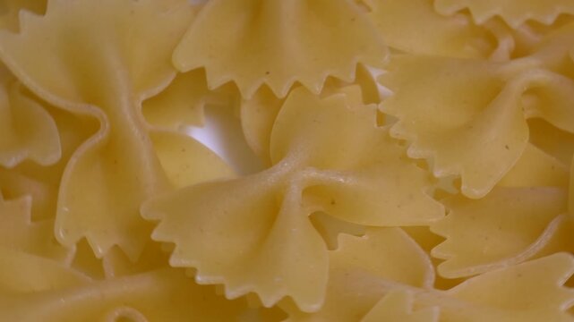 Close-up of raw farfalle bow-tie pasta rotating on a white plate, showcasing its textured shape and golden yellow color under bright studio lighting.