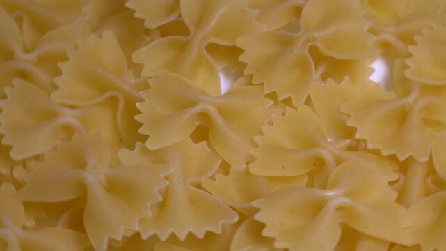 Close-up of raw farfalle bow-tie pasta rotating on a white plate, showcasing its textured shape and golden yellow color under bright studio lighting.