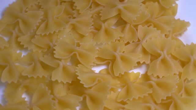 Close-up of raw farfalle bow-tie pasta rotating on a white plate, showcasing its textured shape and golden yellow color under bright studio lighting.