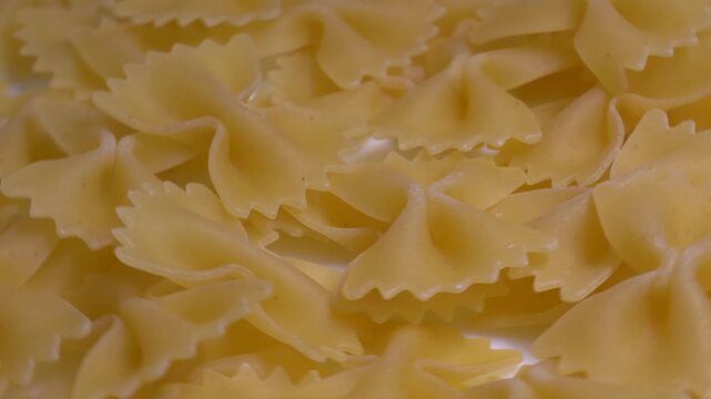 Close-up of raw farfalle bow-tie pasta rotating on a white plate, showcasing its textured shape and golden yellow color under bright studio lighting.