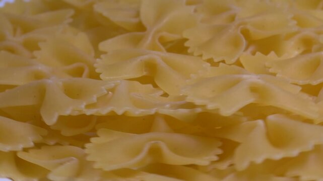 Close-up of raw farfalle bow-tie pasta rotating on a white plate, showcasing its textured shape and golden yellow color under bright studio lighting.
