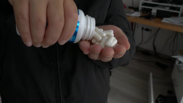 Close-up of a hand pouring white medical capsules from a bottle into an open palm, highlighting the daily health routine under soft lighting.