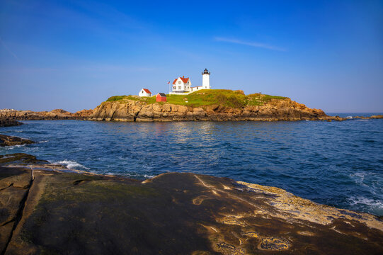 Cape Neddick Nubble Lighthouse on rocky island in York, Maine, with ocean water and smooth rock foreground under clear blue sky