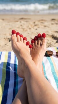 Feet on the beach with raspberries. Women's feet with raspberries on their toes lie on a towel against the background of sand and the sea.