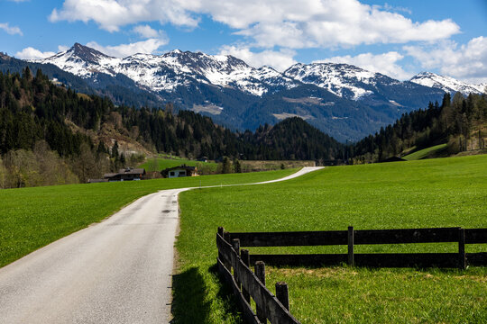 A path leading into the distance and a wooden fence in the spring Tyrolean Windautal valley, in the background snowy Alpine peaks under a blue sky with white clouds