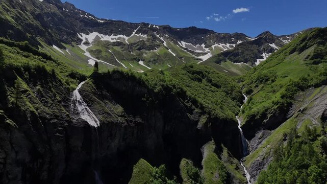 Drone panorama of green alpine slopes with snow patches and waterfalls under a clear blue sky in Arena Batoni, Switzerland.