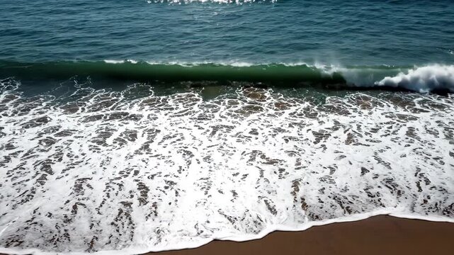 Closeup aerial view of ocean waves crashing onto a sandy beach, creating white foam and ripples on the dark brown shore