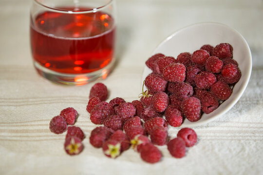 Ripe red raspberries spilled from a white bowl with a blurry glass of juice.
