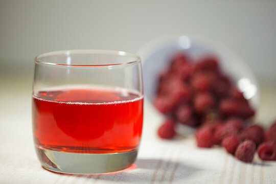Glass of fresh red raspberry juice with blurred berries on a tablecloth background.
