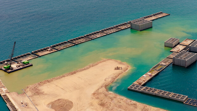 Coastal engineering development shaping new waterfront urban area, featuring modular concrete caissons, dredging, and heavy machinery operating on reclaimed land and barges