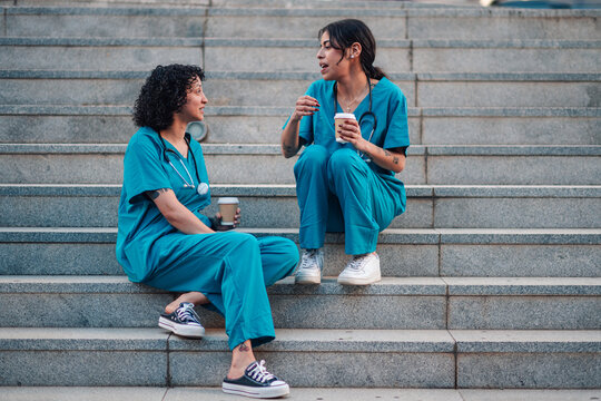 Nurses in scrubs taking coffee break outdoors, talking about work and healthcare, enjoying a moment of friendship