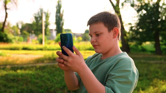 teen boy engrossed in a mobile device, smiling boy using a blue smartphone in warm evening light, casual kid holding a phone with both hands in the park, kid capturing photos on a smartphone in sunny