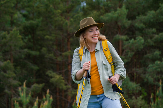 Smiling woman with trekking poles hiking in pine forest with yellow backpack. Concept of Nordic walking, active lifestyle and healthy outdoor sports.