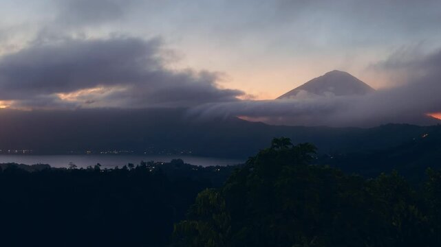 Panoramic View of Mount Abang And Lake Batur at Sunrise, Rolling Clouds Drift Across Mountain Ridge Above Dense Tropical Forest And Calm Water Surface