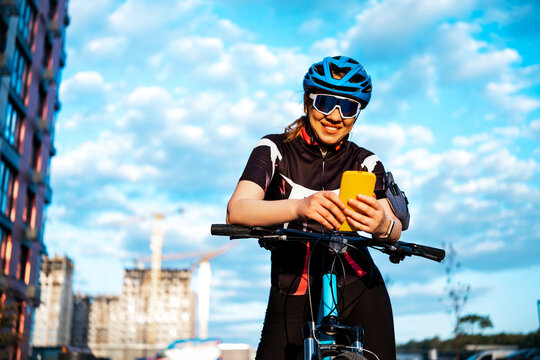 Smiling woman cyclist leaning on bicycle and using smartphone outdoors. Mobile app, active rest concept, modern lifestyle.