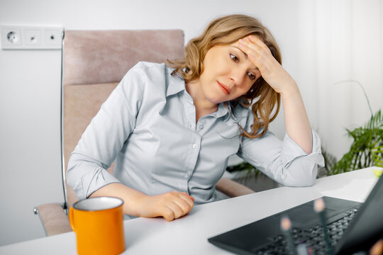 Stressed woman holding her head in pain at laptop desk in office. Workplace stress, headache concept, burnout syndrome.