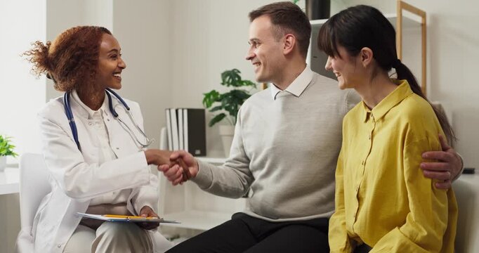 Doctor guides couple in pregnancy planning consultation. Friendly healthcare professional greets happy family in clinic, offering clear advice. Positive family planning healthcare concept.