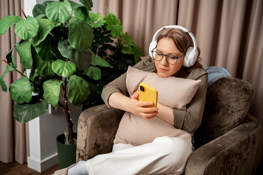 Relaxed woman in headphones hugging pillow holding smartphone in armchair. Home comfort, audio podcast concept, relaxation time.