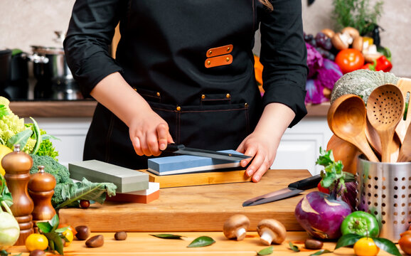 Woman chef sharpening chef knife on whetstone in kitchen with autumn vegetables. Culinary preparation, kitchen knife maintenance, professional culinary skills, autumn comfort food preparation.