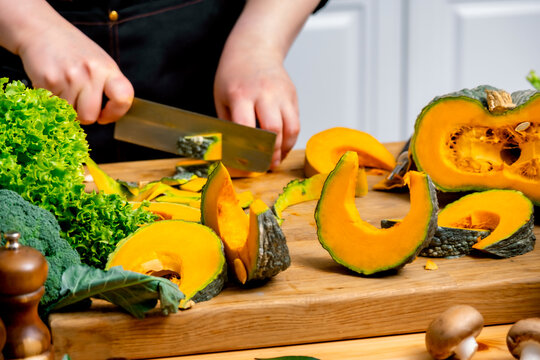 Hands slicing fresh yellow pumpkin on wooden board in modern kitchen. Autumn harvest cooking, healthy comfort food preparation, organic vegetable slicing, culinary prep.