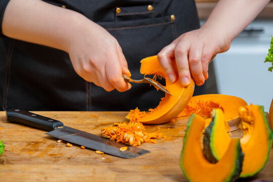 Hands cleaning seeds from pumpkin slice with spoon on wooden board. Autumn harvest prep, raw pumpkin cleaning, healthy home cooking, organic seasonal food concept.