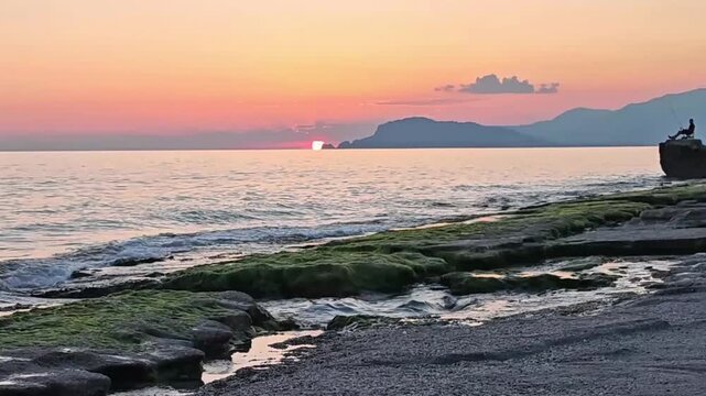 Serene coastal landscape with a fishing man and soft waves hitting mossy rocks at dusk.