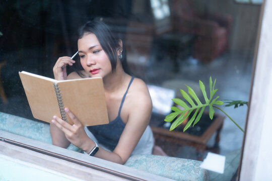 Asian woman sitting on sofa and looking out the window while reading a book, and gazed thoughtfully out of a large glass window in a cozy room with houseplants.