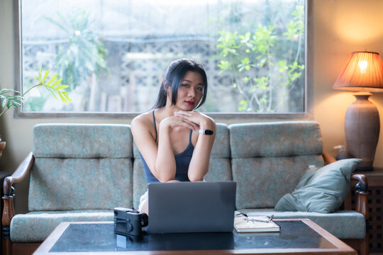Young Asian woman freelancer working in casual wear sitting on a cozy sofa, typing on a laptop computer at a coffee table with a notebook and glasses in a brightly lit living room.