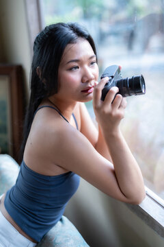 Young Asian female photographer holding a black mirrorless camera, looking thoughtfully out a large glass window in a cozy room with soft daylight.