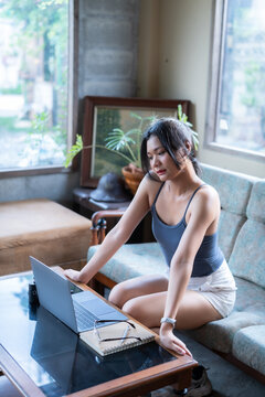 Young Asian woman freelancer working in casual wear sitting on a cozy sofa, typing on a laptop computer at a coffee table with a notebook and glasses in a brightly lit living room.