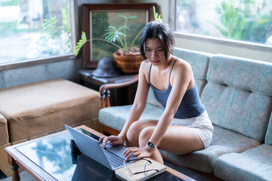 Young Asian woman freelancer working in casual wear sitting on a cozy sofa, typing on a laptop computer at a coffee table with a notebook and glasses in a brightly lit living room.