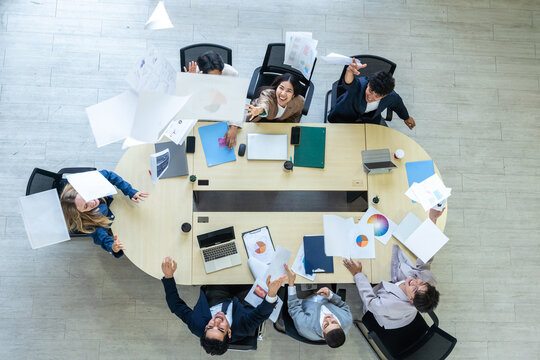 Top view of excited business team around a large conference table, cheering and throwing papers into the air to celebrate successful project completion, freedom, or achievement in a modern office.