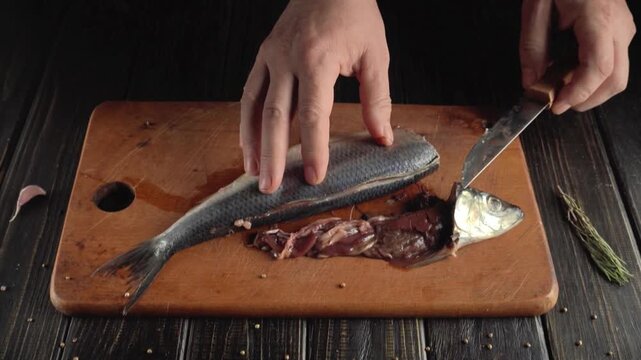 Hands work on a wooden cutting board, cleaning fish by removing internal organs. The kitchen background shows simple tools and spices. The focus is on the fish preparation process