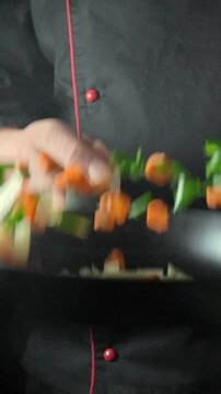 A person is cooking a mix of vegetables in a pan on a stove. The kitchen has steam rising from the pan as the person stirs the ingredients