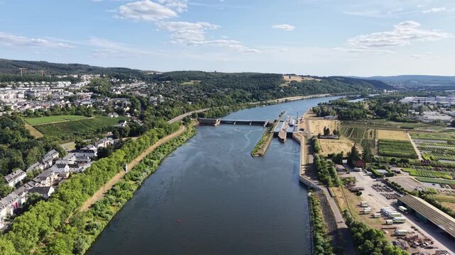 Drone aerial panorama of Trier, Germany, featuring hillside vineyards, the Moselle River, riverside woodland, a navigation lock and residential neighborhoods. Scenic summer landscape with rolling hill