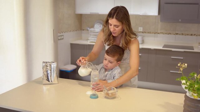 Mother and son preparing baby bottle with milk and cereal in the kitchen