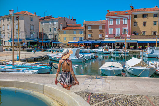 Young woman relaxing by a fountain, overlooking the small boats and colorful Venetian architecture in the historic town of Cres harbor during a sunny summer day of Cres Island in Croatia.