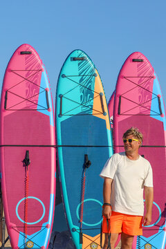 A cheerful man wearing yellow sunglasses, a white t-shirt, and bright orange shorts stands confidently in front of a row of vibrant stand-up paddleboards. 