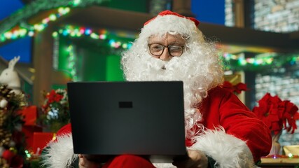 Fototapeta premium Santa Claus makes a video call with children from a cozy Christmas setting after reviewing their gift wish lists on a laptop.