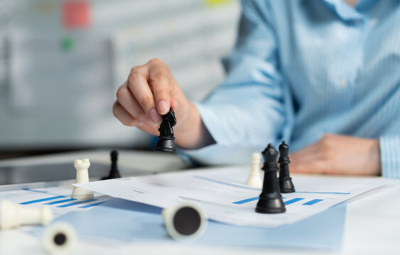 Close up businesswoman hand holding black knight chess piece on financial graph report document. Corporate female planning business strategy and risk management. Leadership and success concept.