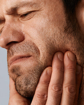 Jaw pain relief closeup of male face and hand pressing cheek muscle, facial tension, stubble, natural skin texture, gray background