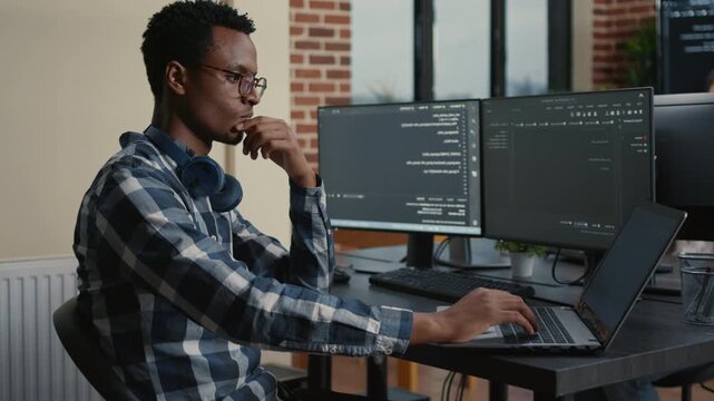 Database developer thinks through a coding problem while touching beard at a laptop desk, reviewing algorithms across multiple screens