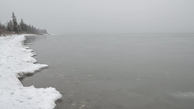 Small waves on a cold looking partially frozen lake as light snow falls on a very gray day
