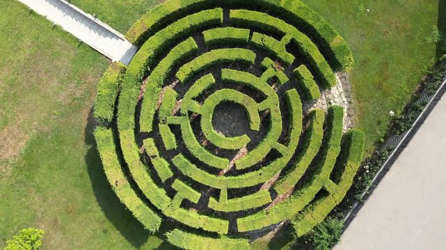 Aerial view shows a circular hedge maze. Lush green hedges form precise concentric paths. Central path leads to an open center. Grassy areas frame the maze